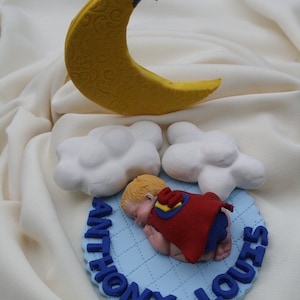 May include: A blue and white cake topper with a sleeping baby wearing a red cape and a yellow crescent moon with a blue star on top. The cake topper says "Anthony Louis" in blue letters.
