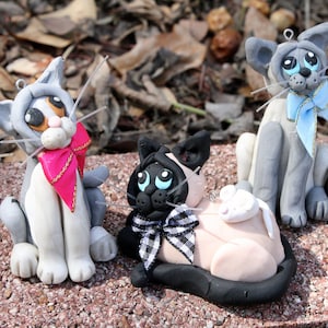 May include: Three handmade ceramic cat figurines. The cat on the left is gray and white with pink and gold ribbon around its neck. The cat in the middle is black and white with a black and white checkered ribbon around its neck. The cat on the right is gray and white with a blue ribbon around its neck.
