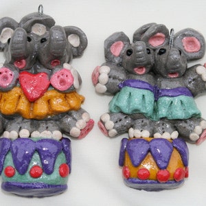 May include: Two whimsical elephant ornaments. Each gray elephant has pink ears and feet, and wears a colorful outfit. One has an orange skirt and a red heart, the other has a teal skirt. Both have a purple drum.