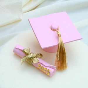 May include: A pink graduation cap with a gold tassel and a rolled-up scroll tied with a gold ribbon. The scroll has the year "2025" printed on it. The items are on a white surface with a soft, white fabric background.