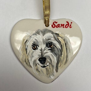 May include: White ceramic heart ornament featuring a hand-painted portrait of a small, white dog with grey markings. The dog's name, "Sandi", is written in red lettering on the ornament.