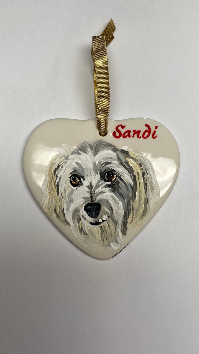 May include: White ceramic heart ornament featuring a hand-painted portrait of a small, white dog with grey markings. The dog's name, "Sandi", is written in red lettering on the ornament.