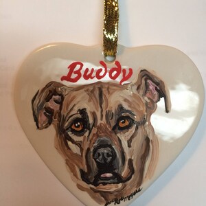 May include: A white heart-shaped ceramic ornament featuring a painted portrait of a brown dog. The dog's name, "Buddy", is written in red letters above the dog's head.