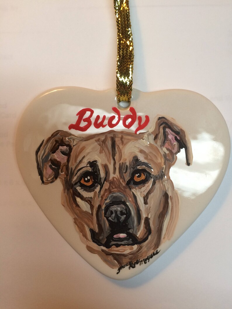 May include: A white heart-shaped ceramic ornament featuring a painted portrait of a brown dog. The dog's name, "Buddy", is written in red letters above the dog's head.