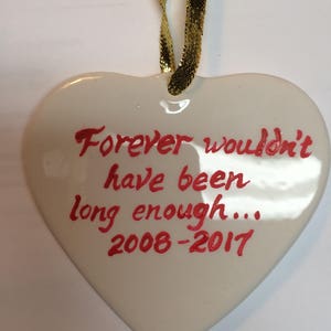 May include: White ceramic heart ornament with a gold ribbon. The heart has red handwritten text that reads "Forever wouldn't have been long enough... 2008-2017".