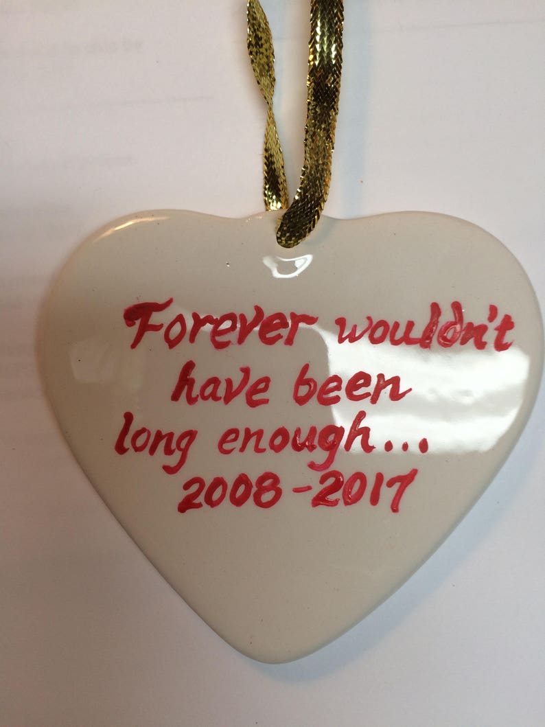 May include: White ceramic heart ornament with a gold ribbon. The heart has red handwritten text that reads "Forever wouldn't have been long enough... 2008-2017".
