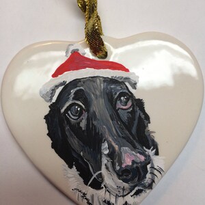 May include: A hand-painted ceramic heart ornament featuring a black and white dog wearing a red Santa hat. The ornament has a gold ribbon for hanging.