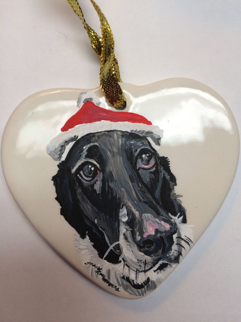 May include: A hand-painted ceramic heart ornament featuring a black and white dog wearing a red Santa hat. The ornament has a gold ribbon for hanging.