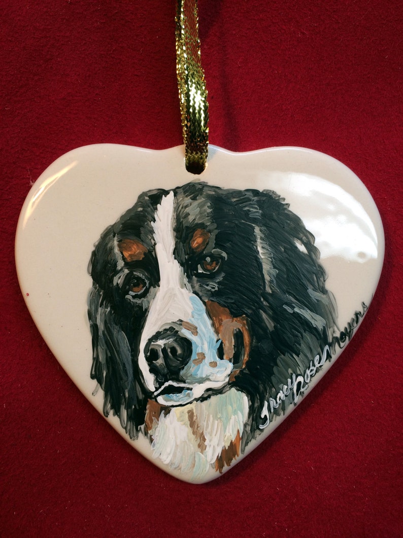 May include: A white ceramic heart-shaped ornament featuring a hand-painted portrait of a Bernese Mountain Dog. The dog's coat is black, white and brown. The ornament is signed "Tracy Rosen Myers".