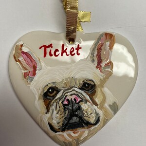 May include: A white ceramic heart-shaped ornament featuring a hand-painted portrait of a French Bulldog. The dog has brown eyes, a black nose, and a pink tongue. The ornament is decorated with a gold ribbon and the name "Ticket" is written in red paint.