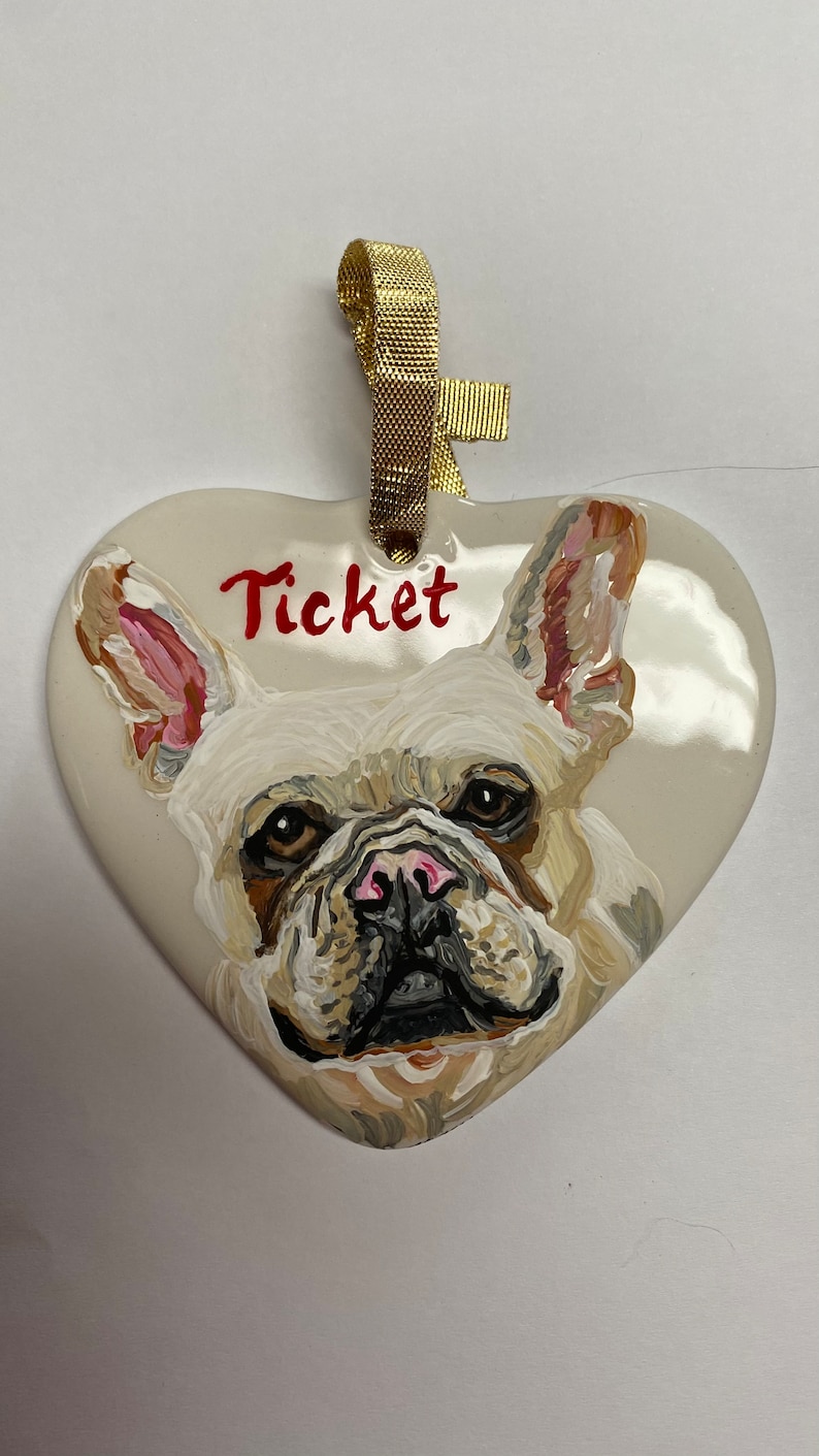 May include: A white ceramic heart-shaped ornament featuring a hand-painted portrait of a French Bulldog. The dog has brown eyes, a black nose, and a pink tongue. The ornament is decorated with a gold ribbon and the name "Ticket" is written in red paint.