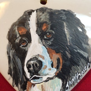 May include: Hand-painted ceramic heart ornament featuring a close-up portrait of a Bernese Mountain Dog with black, white and brown fur. The ornament has a gold ribbon attached for hanging and is signed "Grace Rosen".