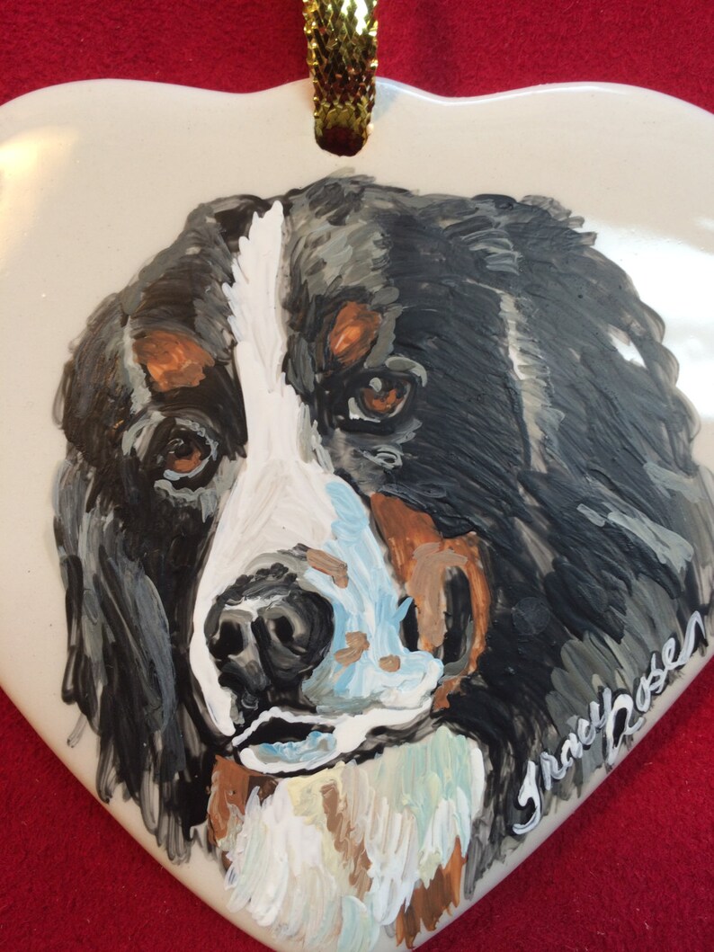May include: Hand-painted ceramic heart ornament featuring a close-up portrait of a Bernese Mountain Dog with black, white and brown fur. The ornament has a gold ribbon attached for hanging and is signed "Grace Rosen".