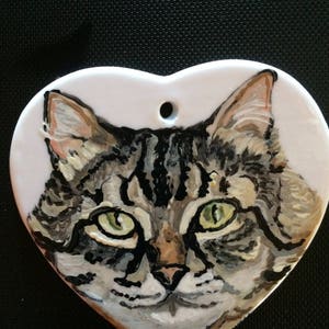 May include: A hand-painted ceramic heart ornament featuring a close-up portrait of a tabby cat with green eyes. The cat's fur is depicted in shades of brown, black, and white.