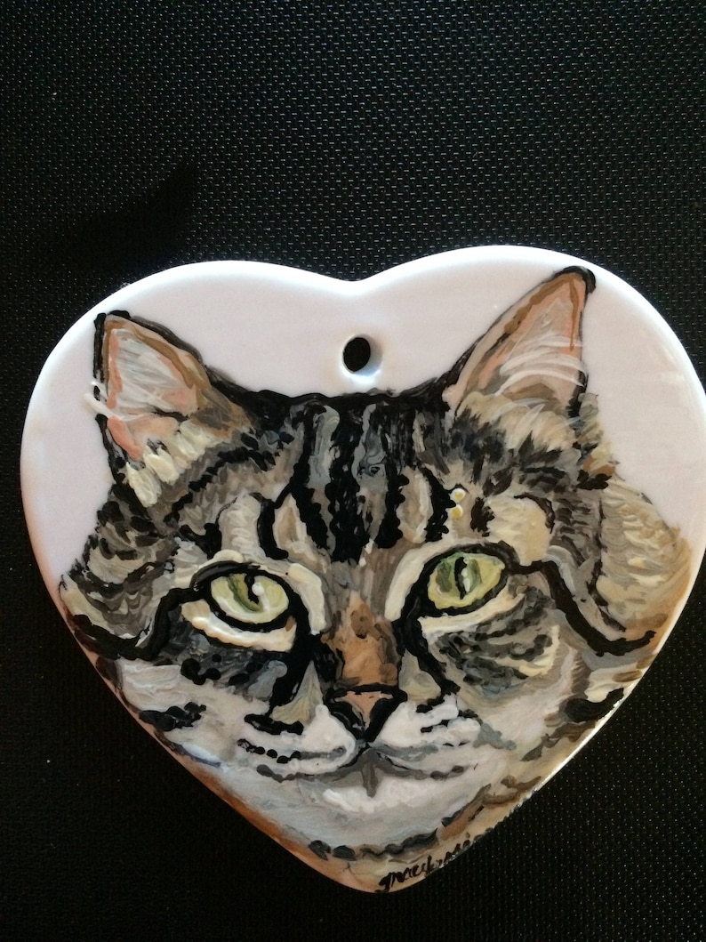 May include: A hand-painted ceramic heart ornament featuring a close-up portrait of a tabby cat with green eyes. The cat's fur is depicted in shades of brown, black, and white.