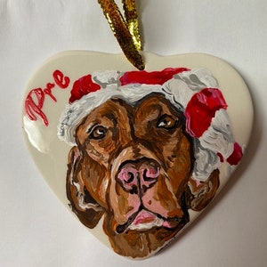 May include: A heart-shaped ceramic ornament featuring a painted image of a brown dog wearing a red and white Santa hat. The ornament is inscribed with the name "Pre" in red paint.