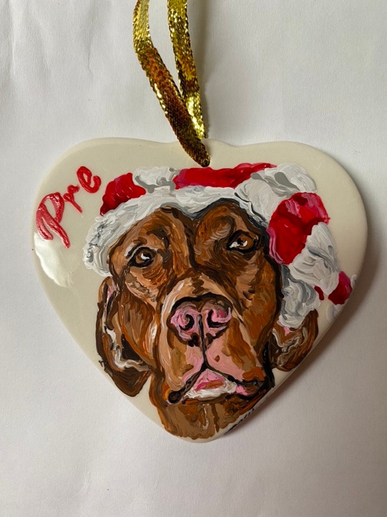 May include: A heart-shaped ceramic ornament featuring a painted image of a brown dog wearing a red and white Santa hat. The ornament is inscribed with the name "Pre" in red paint.