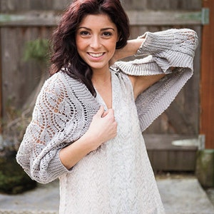 May include: A woman wearing a white top with a grey crocheted cardigan. She is smiling and has long brown hair.