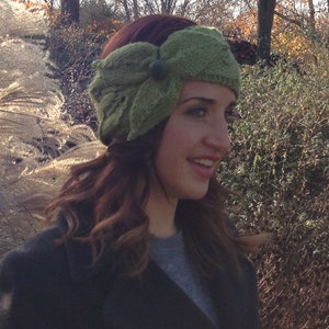 May include: A green knitted headband with a leaf design and a small button detail. The headband is worn by a person with auburn hair and a dark coat. The background features trees and foliage.