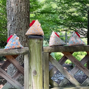 May include: Four triangular fabric objects with red accents and floral patterns rest on a wooden fence. The fabrics feature orange, blue, and white floral designs. The objects are set against a backdrop of green trees and foliage.