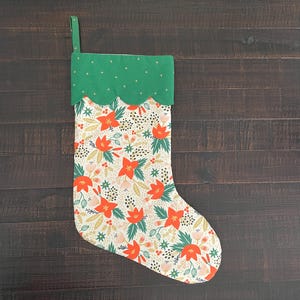 May include: A fabric Christmas stocking featuring a cream-colored body with a repeating floral pattern of red flowers, green leaves, and small berries. The top cuff is solid green with a scalloped bottom edge and a small fabric hanging loop.