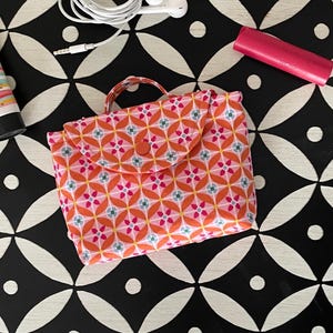 May include: A small, patterned fabric pouch with a button closure, featuring a repeating floral design in shades of orange, pink, and blue. The pouch has a small handle and is set against a black and white geometric background. Also visible are a tube of lotion, earbuds, and a pink lip balm.