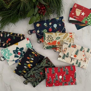 May include: A collection of small fabric wallets featuring various holiday-themed patterns including Christmas lights, ornaments, pine trees, and holly. Each rectangular wallet has a snap closure and a metal D-ring attached to the side for easy carrying.