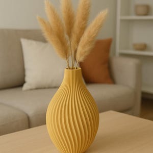 Puede incluir: Un jarrón amarillo mostaza acanalado que contiene varios tallos beige secos y plumosos. El jarrón está sobre una mesa de madera clara. El fondo incluye un sofá beige, cojines y una estantería blanca.