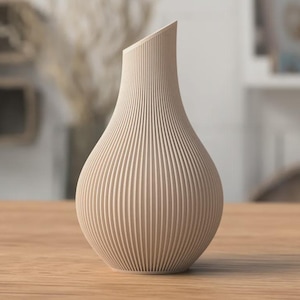 Può includere: Un vaso beige a coste con una parte superiore inclinata. Il vaso ha una base bulbosa e un collo sottile, posto su una superficie di legno. Il design presenta linee verticali, creando un aspetto strutturato. Il vaso è un elemento decorativo per la casa.