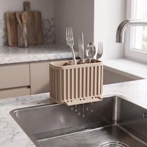 Può includere: Uno scolaposate beige, rettangolare e scanalato, con molteplici scomparti interni, posizionato sul bordo di un lavello in acciaio inossidabile. Lo scolaposate presenta piccoli fori di drenaggio alla base per far defluire l'acqua.