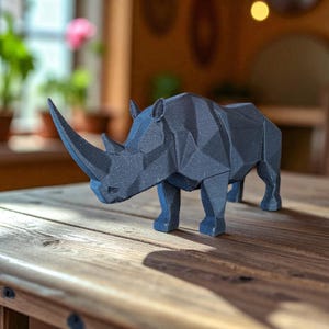 Puede incluir: Una escultura de rinoceronte geométrico gris oscuro con un cuerno largo, de pie sobre una superficie de madera. El rinoceronte está hecho de formas angulares, lo que le da un aspecto moderno y artístico. Es un objeto decorativo.