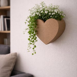 Könnte beinhalten: Ein herzförmiger Wandtopf aus hellbraunem Material, gefüllt mit grünen Hängepflanzen und kleinen weißen Blüten. Der Topf ist an einer weißen Wand befestigt, im Hintergrund sind ein Sofa und Regale zu sehen.