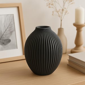 Könnte beinhalten: Eine schwarze, gerippte Keramikvase mit runder Form. Die Vase ist etwa 20 cm hoch und steht auf einer hellen Holzoberfläche. Im Hintergrund befinden sich weitere dekorative Gegenstände, darunter ein gerahmter Druck und eine Kerze.