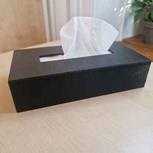 Puede incluir: Una caja rectangular para pañuelos con un acabado negro mate texturizado. La caja presenta una abertura rectangular central en la parte superior, a través de la cual se extrae un pañuelo de papel blanco que sobresale ligeramente.