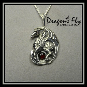 Op de afbeelding: Zilveren drakenhanger met een rode edelsteen, aan een zilveren ketting. De hanger is in de vorm van een draak die om een rode edelsteen is gekruld. De hanger hangt aan een zilveren ketting. De hanger is gemarkeerd met "Dragons Fly Designs".