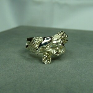 May include: A silver ring with a detailed design of two intertwined figures. The ring features a small, clear gemstone.