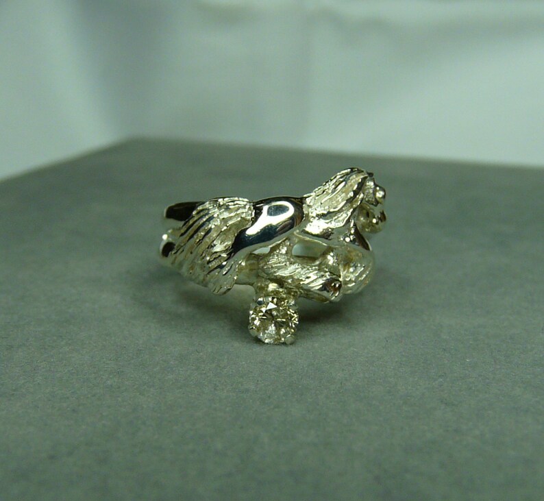 May include: A silver ring with a detailed design of two intertwined figures. The ring features a small, clear gemstone.