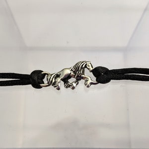 May include: A black cord bracelet with a silver horse charm. The horse is running and has a detailed mane.