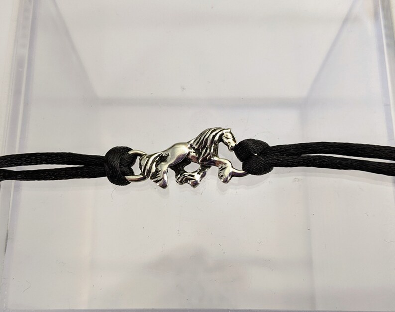 May include: A black cord bracelet with a silver horse charm. The horse is running and has a detailed mane.