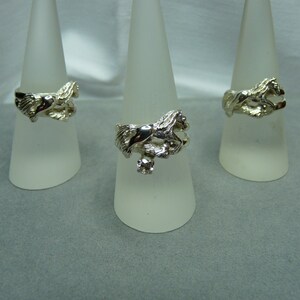 May include: Three silver rings with a horse design. The rings are displayed on white cone-shaped stands.