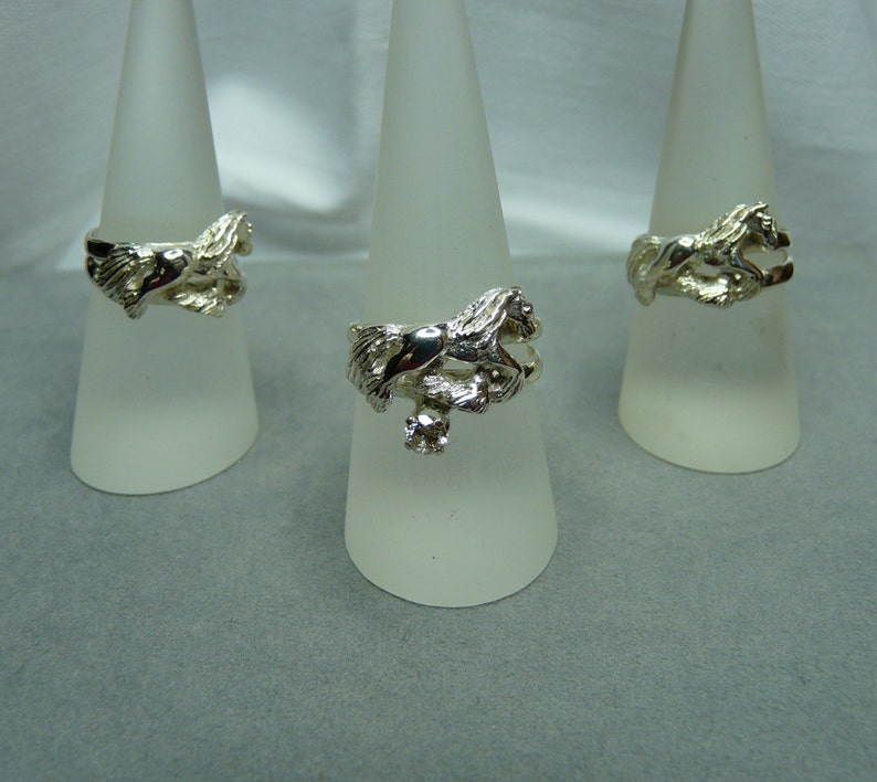 May include: Three silver rings with a horse design. The rings are displayed on white cone-shaped stands.