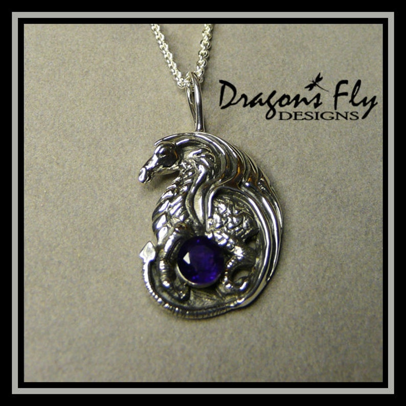Op de afbeelding: Sterling zilveren hanger met een gedetailleerd drakenontwerp met een paarse amethist edelsteen. De hanger is aan een zilveren ketting en heeft de tekst "Dragons Fly Designs" op de achtergrond.