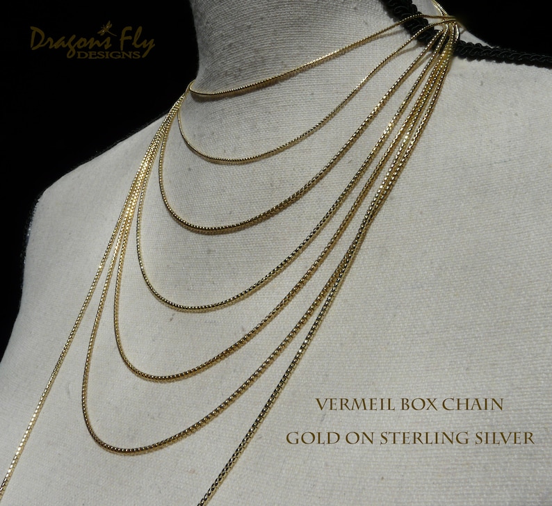 May include: A set of gold vermeil box chains of varying lengths, displayed on a white mannequin. The chains are made of gold on sterling silver. The text "VERMEIL BOX CHAIN GOLD ON STERLING SILVER" is displayed at the bottom of the image.
