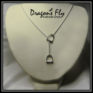 May include: A silver necklace with two dangling stirrup pendants. The necklace is on a white display stand. The text "Dragon's Fly DESIGNS" is visible in the background.