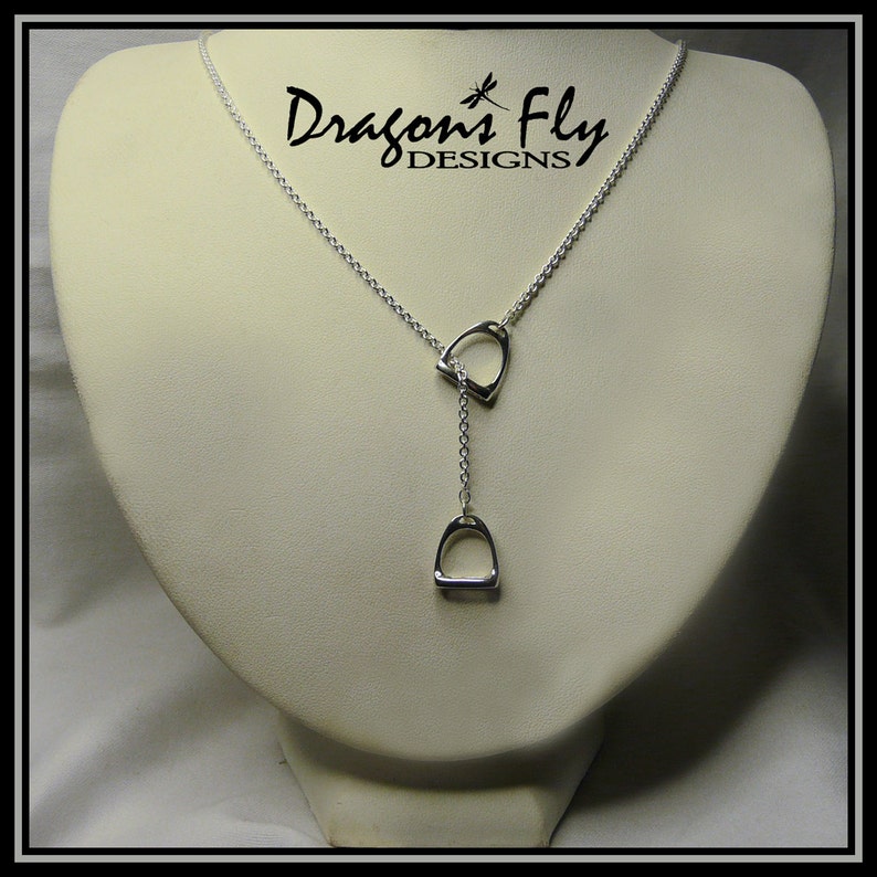 May include: A silver necklace with two dangling stirrup pendants. The necklace is on a white display stand. The text "Dragon's Fly DESIGNS" is visible in the background.