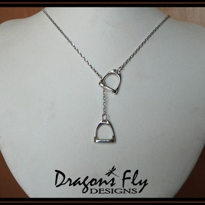 May include: Silver necklace with two dangling stirrup pendants. The necklace is on a white display stand. The text "Dragon's Fly DESIGNS" is visible at the bottom of the image.
