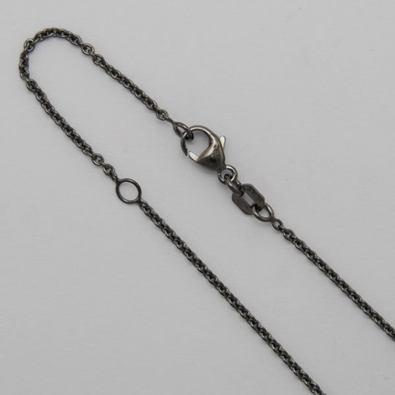 K&ouml;nnte beinhalten: Nahaufnahme einer anthrazitfarbenen Halskette mit einem Karabinerverschluss und einem verstellbaren Ring. Die Kette besteht aus kleinen, miteinander verbundenen Gliedern, die ein zartes, aber dennoch haltbares Design ergeben. Die Halskette wird vor einem schlichten, hellen Hintergrund pr&auml;sentiert.