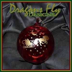 May include: A red glass Christmas ornament with a gold design of Santa Claus in his sleigh pulled by reindeer flying through a starry night sky. The ornament has a gold crescent moon and stars.