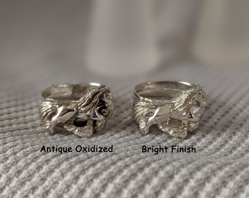 May include: Two silver rings with a detailed design of a horse running. The ring on the left has an antique oxidized finish, while the ring on the right has a bright finish. The rings are displayed on a gray fabric background.