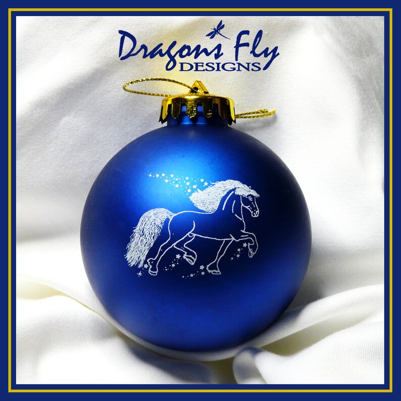 May include: A blue Christmas ornament with a white silhouette of a horse running with a star trail behind it. The ornament is decorated with gold trim and has the text "Dragons Fly Designs" on the top.